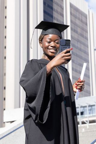African american graduate taking selfie in cap and gown on urban steps with diploma