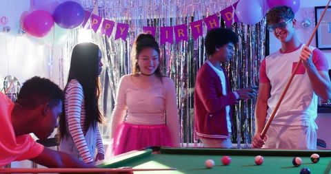 Multicultural young adults playing pool at birthday party with pink tutu and balloons
