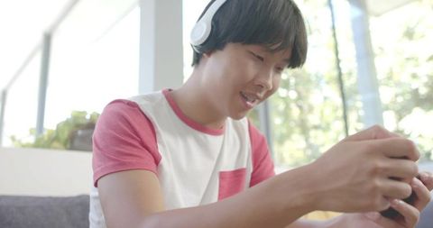 Teenager wearing headphones playing smartphone games on couch in bright sunlit living room