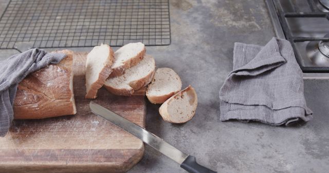 This image shows freshly baked bread sliced on a rustic cutting board with a knife nearby. It is set on a kitchen counter with a gray linen cloth, providing a homely and cozy atmosphere. Ideal for use in bakery marketing materials, food blogs, cooking websites, or home culinary articles.