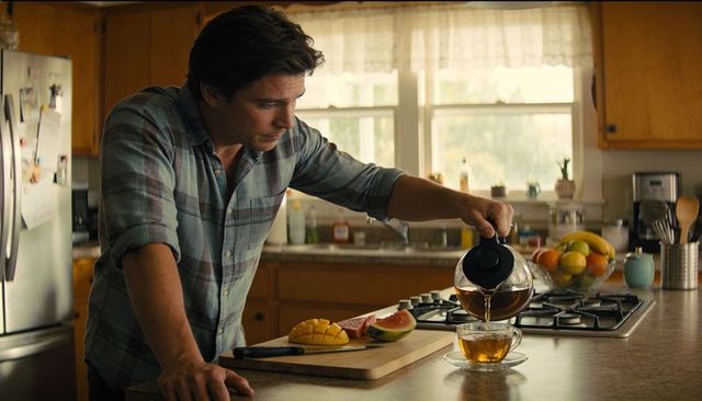 Middle-aged man pouring tea at sunlit kitchen island with fresh mango and fruit bowl