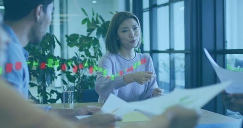 Asian woman leading team meeting in glass office pointing at documents with stock chart overlay