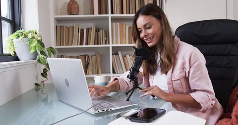 Smiling woman podcasting at home office desk with laptop and microphone