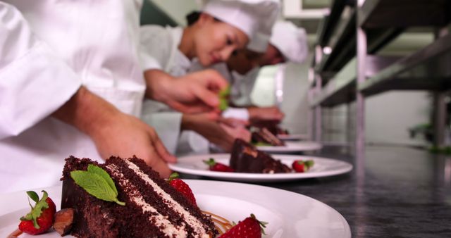 Chefs in white uniforms are focusing on meticulously preparing and garnishing dessert plates in a professional kitchen. A close-up emphasizes a chocolate cake with strawberries and mint leaves being artistically set on white plates. Ideal for use in content related to culinary arts, gourmet cooking, restaurants, baking techniques, and team collaboration in professional kitchens.