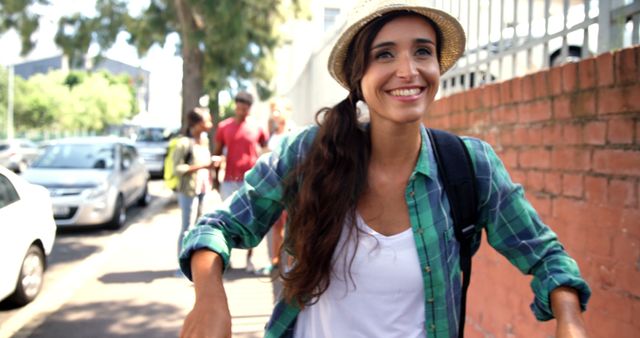 Woman enjoys walking on a sunny urban sidewalk, dressed in casual attire with a green plaid shirt and straw hat. Ideal for concepts related to travel, summer, joy, and city life. Useful for advertisements, travel blogs, lifestyle articles, and social media campaigns.