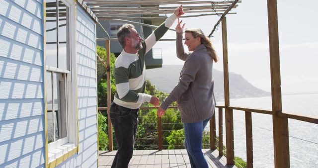 Senior couple dancing happily on balcony overlooking ocean and mountains. Perfect for concepts of retirement happiness, active senior lifestyle, outdoor leisure activities, couples' joy, and healthy aging.