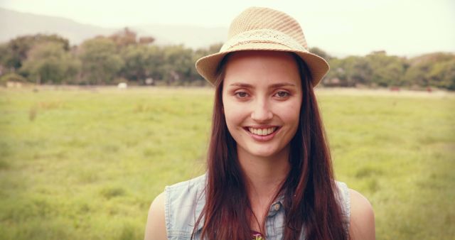 A young Caucasian woman smiles gently, wearing a straw hat in a serene outdoor setting, with copy space. Her casual attire and the natural backdrop suggest a relaxed, leisurely day.