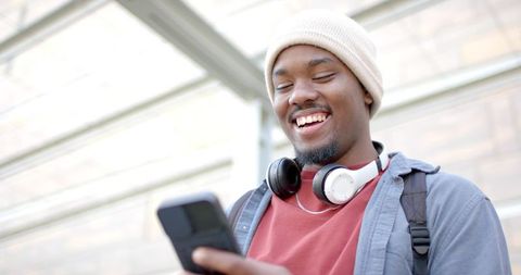 Smiling african american man checking phone with headphones and backpack in urban daylight
