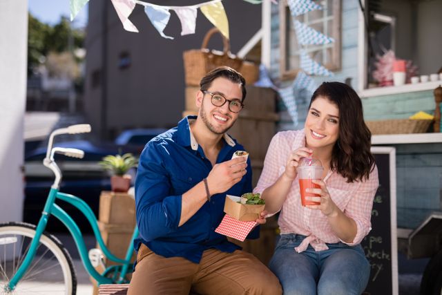 Young couple sitting outdoors, enjoying snacks and juice. They are smiling and appear relaxed, suggesting a casual and happy moment. Ideal for use in lifestyle, food and drink, summer, and leisure-related content.
