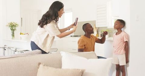 Mother capturing son's height while father measuring and encouraging in bright kitchen
