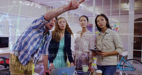 Leader pointing while team collaborating around laptop and tablet in glass-walled office