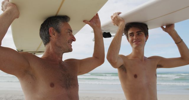 Father and son carrying surfboards, smiling and walking along a sandy beach toward the ocean on a sunny day. This image can be used to depict family activities, surfing culture, beach vacation promotions, or active lifestyle advertisements. Great for websites, travel blogs, and marketing materials focused on family, adventure, and outdoor sports.