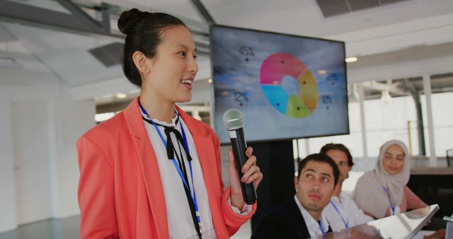 Side view close up of a young Asian businesswoman wearing a pink jacket standing at a lectern using a microphone to address the audience at a business conference. In the background is a screen with information on it and a diverse group of business people sitting, looking up at her and listening