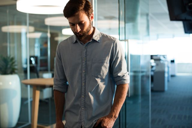 Businessman standing in modern office with glass walls, looking down pensively. Ideal for use in articles about workplace stress, corporate challenges, mental health in the workplace, or professional introspection.