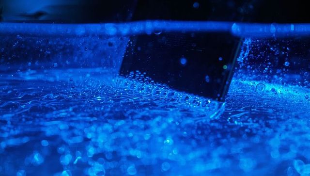 Black smartphone half-submerged in blue-lit water with bubbling ripples and reflections