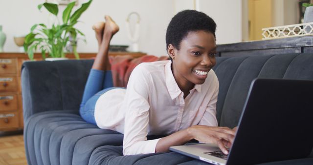 Woman lying on blue couch, using laptop with smile. Perfect for illustrating remote work, leisure at home, and online activities. Suitable for websites, blogs, ads related to home living, technology, and lifestyle.