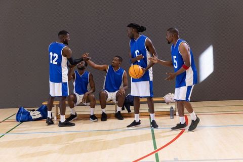 African american basketball team discussing strategy on bench holding ball in indoor gym