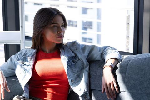 Contemplative woman relaxing on highrise sofa in red top and denim jacket with smartwatch