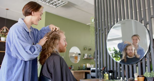 Smiling woman styling long hair of red-bearded man in modern hair salon, with green and wood interiors. Perfect for content related to personal grooming, hairdressing services, beauty industry promotions, and modern salon design inspiration.