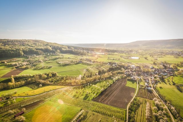 Aerial shot capturing the peaceful ambiance of a rural farming village at sunrise. The fields are lush green, dotted with plowed sections ready for planting. Ideal for use in travel brochures, countryside living magazines, agricultural websites, and environmental conservation promotions.
