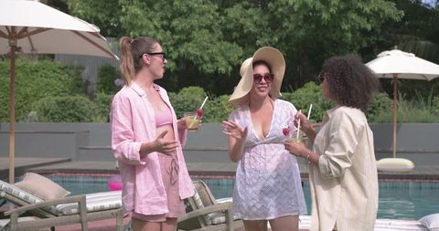 Multicultural women laughing and chatting poolside in cover-ups, sipping cocktails