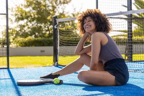 African american woman smiling and resting on blue paddle court in sunny outdoor scene