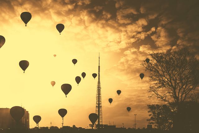 Silhouetted hot air balloons floating across the sunset sky create a serene and adventurous atmosphere. The radio tower stands tall amidst the calm scene, adding contrast to the expansive sky. Ideal for use in themes promoting travel, peace, and exploration.