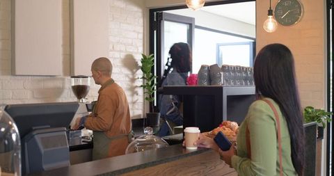 Customer waiting holding coffee and phone while barista preparing espresso in industrial cafe