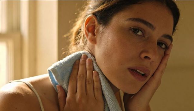 Hispanic woman wiping face with towel by window wearing thin-strap top intimate self-care