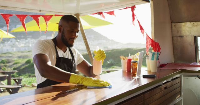 African american man wearing apron and gloves cleaning the food truck with disinfectant spray. hygiene and safety during coronavirus covid 19 pandemic concept