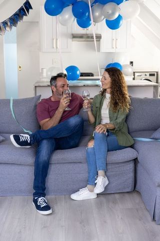 Middle-aged couple toasting wine on sofa during blue silver balloon birthday celebration