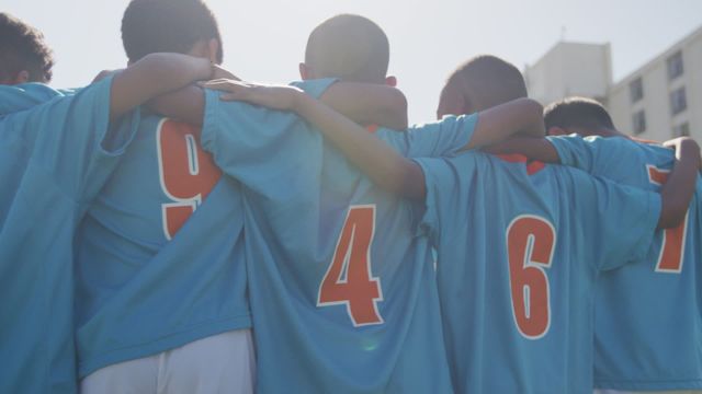 Boys from multi-ethnic soccer teams stand together embracing, showcasing teamwork and unity on a sunny outdoor field. Their blue jerseys with numbers highlight their team spirit, while the sunny glow emphasizes a sense of warmth and camaraderie. Perfect for themes focused on teamwork, child development through sports, diversity, and inclusion. Excellent for educational materials, sports club promotions, and motivational imagery.