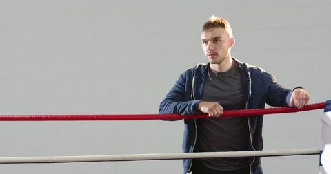 Contemplative boxer leaning on red ring rope in minimalist gym with dramatic lighting