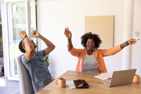 African american couple celebrating win at home workspace with laptop and paperwork