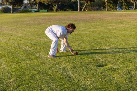 Sunlit athlete wearing white sportswear bending to pick up red ball on green sports field
