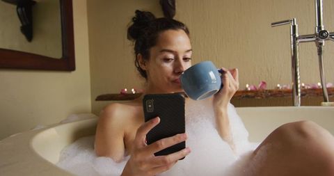 Young woman relaxing in bubble bath with coffee and smartphone
