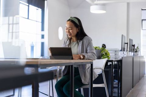 Asian woman working on tablet at modern coworking desk, smiling and focused