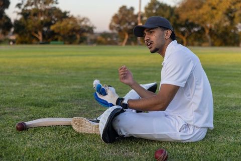 Indian cricketer sitting on grass adjusting batting gloves beside bat during golden hour