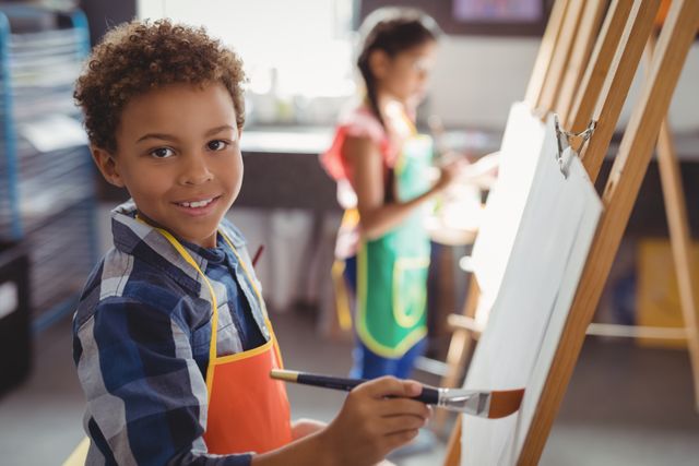 Schoolboy smiling while painting on canvas in classroom. Ideal for educational content, art class promotions, creative learning materials, and children's activities. Highlights creativity, learning, and joy in education.