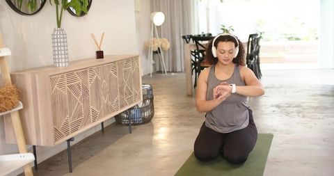 African american woman checking smartwatch while kneeling on yoga mat wearing headphones