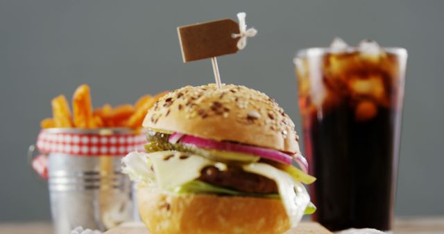 A delicious burger with a variety of toppings is presented in the foreground, accompanied by a side of fries and a cold soda in the background. The setup suggests a casual dining experience or a quick service restaurant offering.