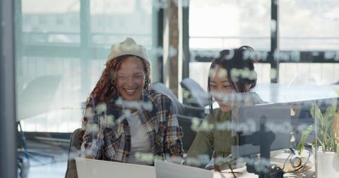 Laughing diverse female coworkers collaborating at laptop behind glass with sticky notes