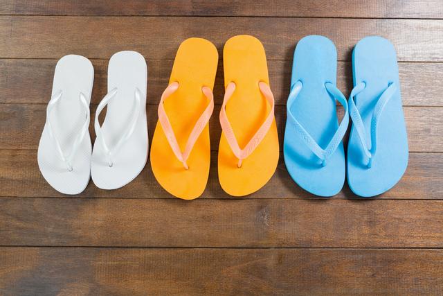 Three pairs of flip flops in white, orange, and blue are neatly arranged on a wooden floor. This image is perfect for summer-themed promotions, travel blogs, vacation advertisements, and casual footwear marketing.