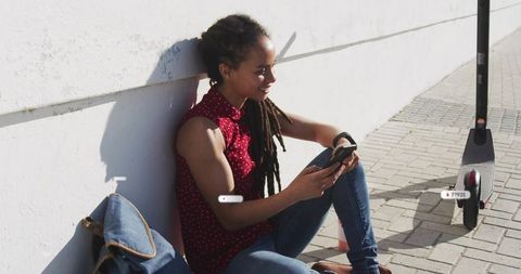 Woman sitting sidewalk with smartphone and scooter outdoors