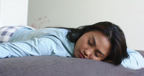 Asian woman lying face down on grey sofa resting in blue top and checkered pajama bottoms