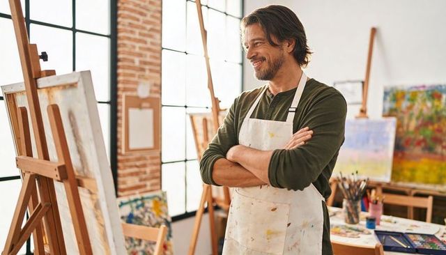 Confident painter standing in sunlit loft studio wearing paint-stained apron and smiling