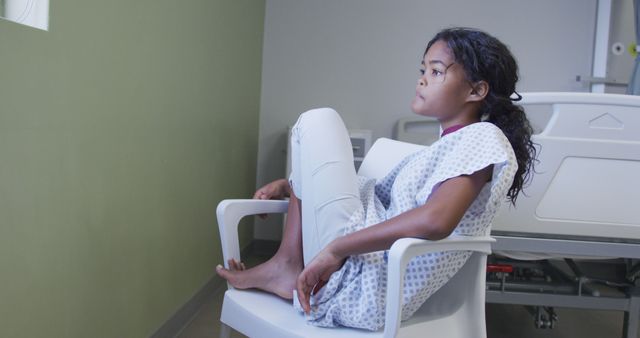 Pensive girl sitting in a white chair wearing a hospital gown. Ideal for themes related to healthcare, patient emotions, hospital interiors, child health concerns, medical illustrations, and mental health.