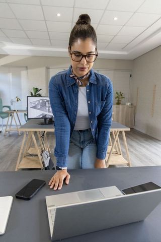 Young female architect leaning over laptop reviewing floor plans in modern design studio