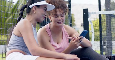 African american women sharing phone on tennis court bench, resting after workout