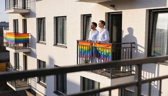 Gay couple standing on apartment balcony wearing matching pajamas with rainbow pride flags